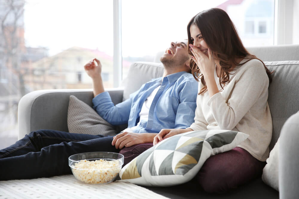 man and woman laughing and eating popcorn on a settee